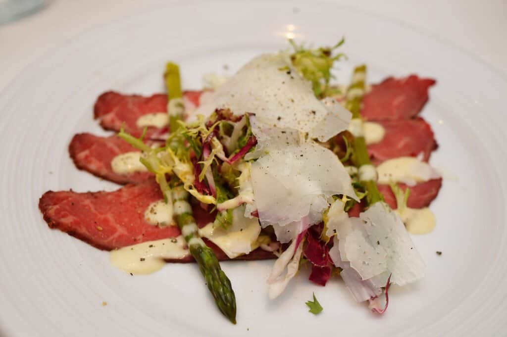 Beef carpaccio topped with microgreens, shaved Parmesan, and drizzled with creamy dressing, served o.