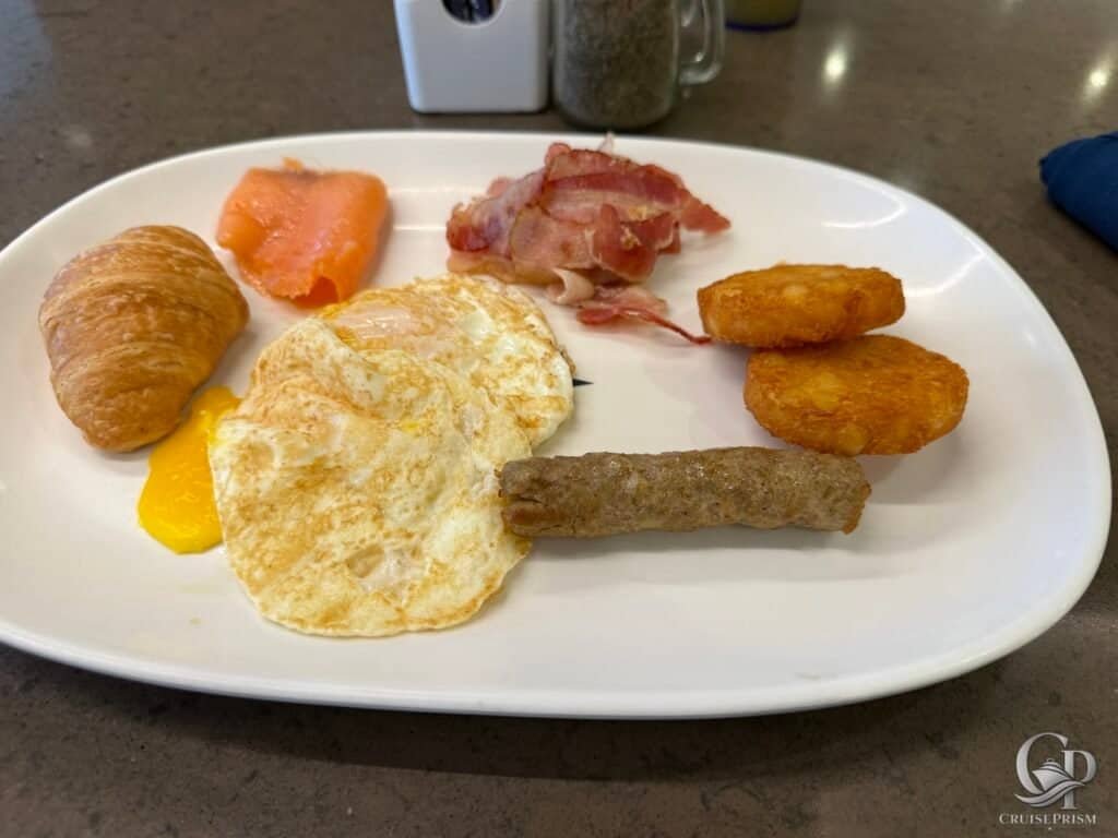 Egg, bacon, hash brown, salmon, croissant, and fried potato wedges on a white plate.